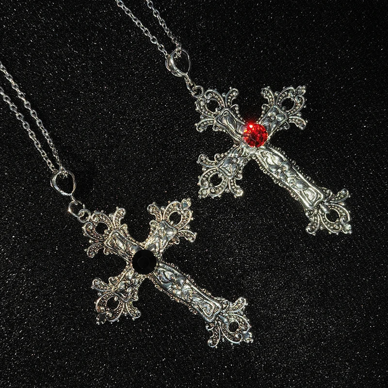 Two silver gothic cross pendant necklaces, one with a red stone and one with a black stone, on a dark background